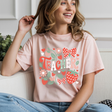 Woman wearing a pink t-shirt with 'Teacher' and floral designs, sitting on a couch.