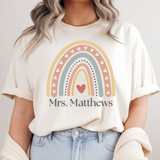 Person wearing a white t-shirt with a rainbow and heart design, text 'Mrs. Matthews', on a plain background.