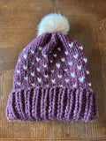 Purple knitted hat with white heart pattern and white pom-pom on a wooden surface