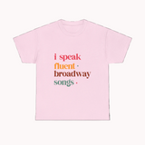 Pink t-shirt with colorful text 'i speak fluent broadway songs' on a white background