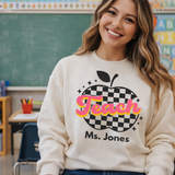 Teacher wearing a sweatshirt with 'Teach' and an apple design in a classroom setting
