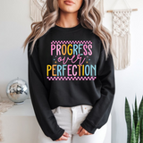 Person wearing a black sweatshirt with 'Progress over Perfection' text in colorful letters, standing in a room with decor elements.